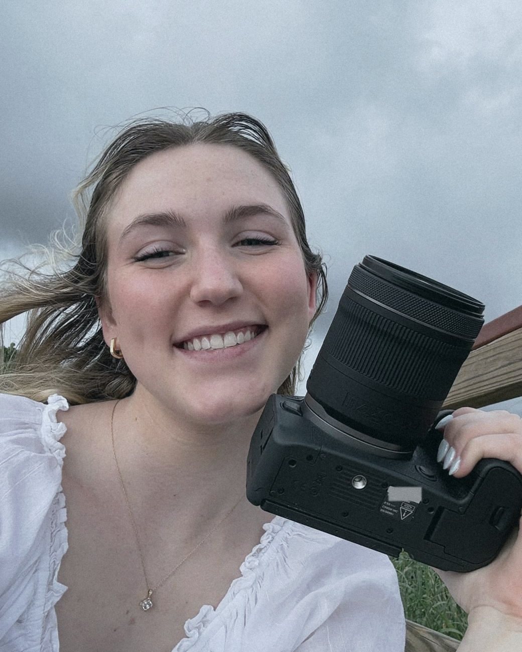 Aurelia Dougan in a white ruffled blouse, smiling at the camera and holding her Canon beside her face, with a wooden railing behind her.